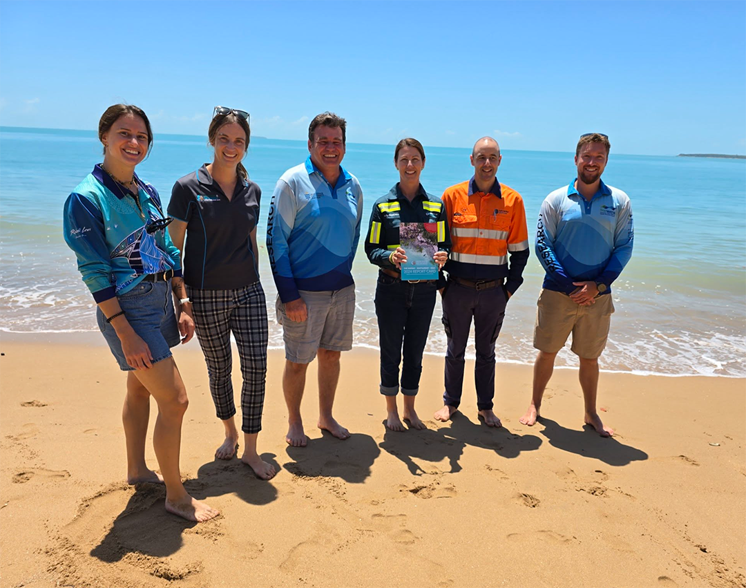 sip-funders-jcu A group of people stand on a beach with the ocean in the background. One of the them is holding the Report Card.