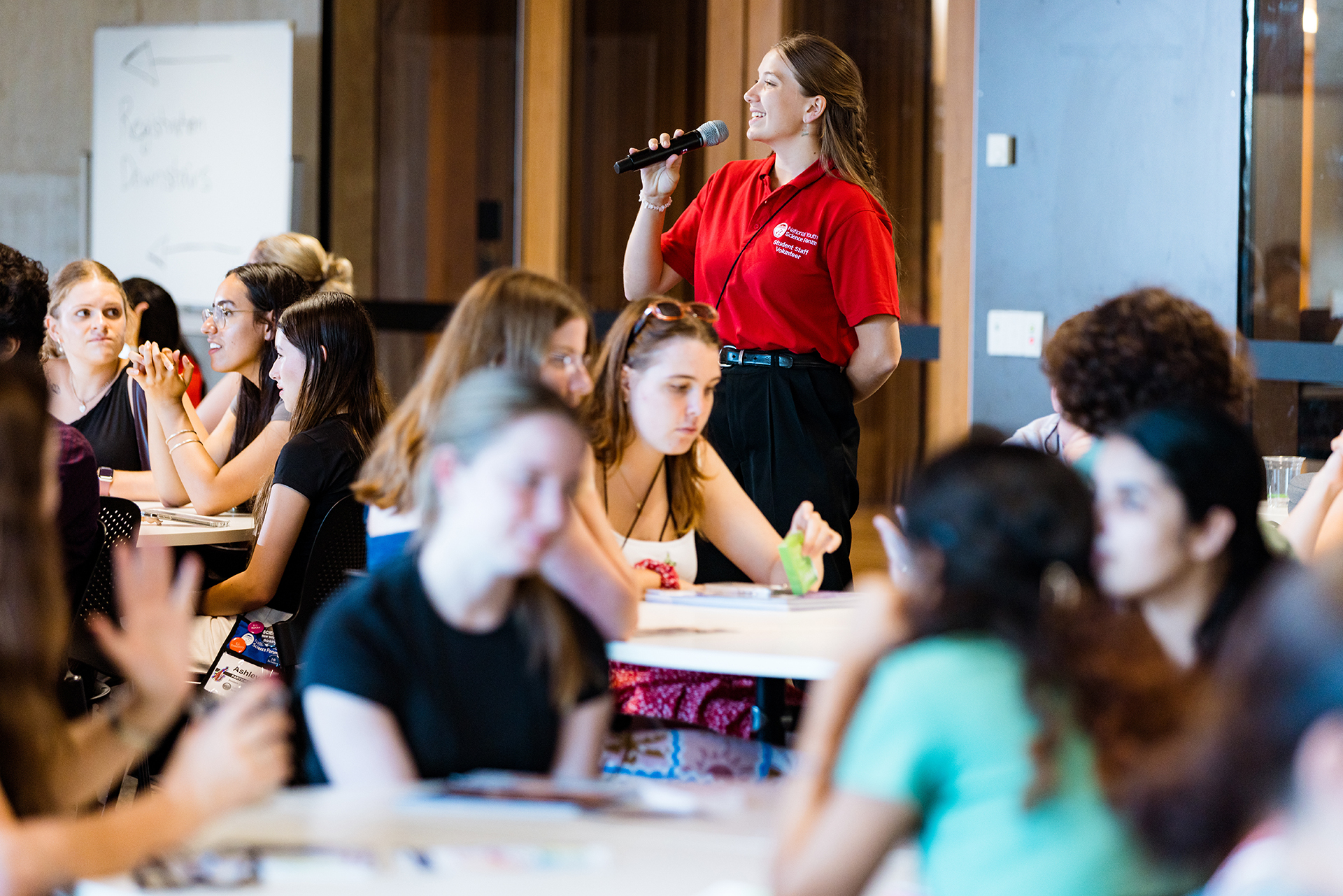 A woman stands at the front of a room holding a microphone. Young students are sitting at tables in the foreground.