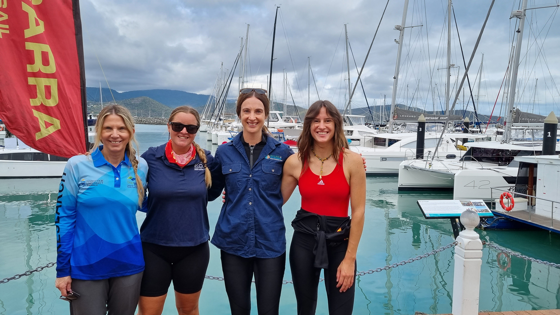 Four women standing on a dock in front of boats in the water.