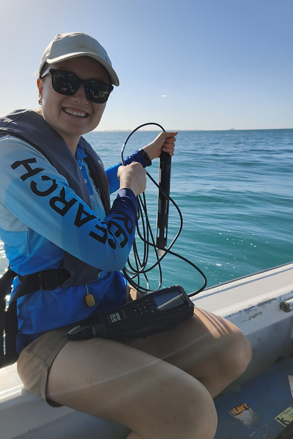 Woman sits on the edge of a boat lowering water quality testing equipment into the water.